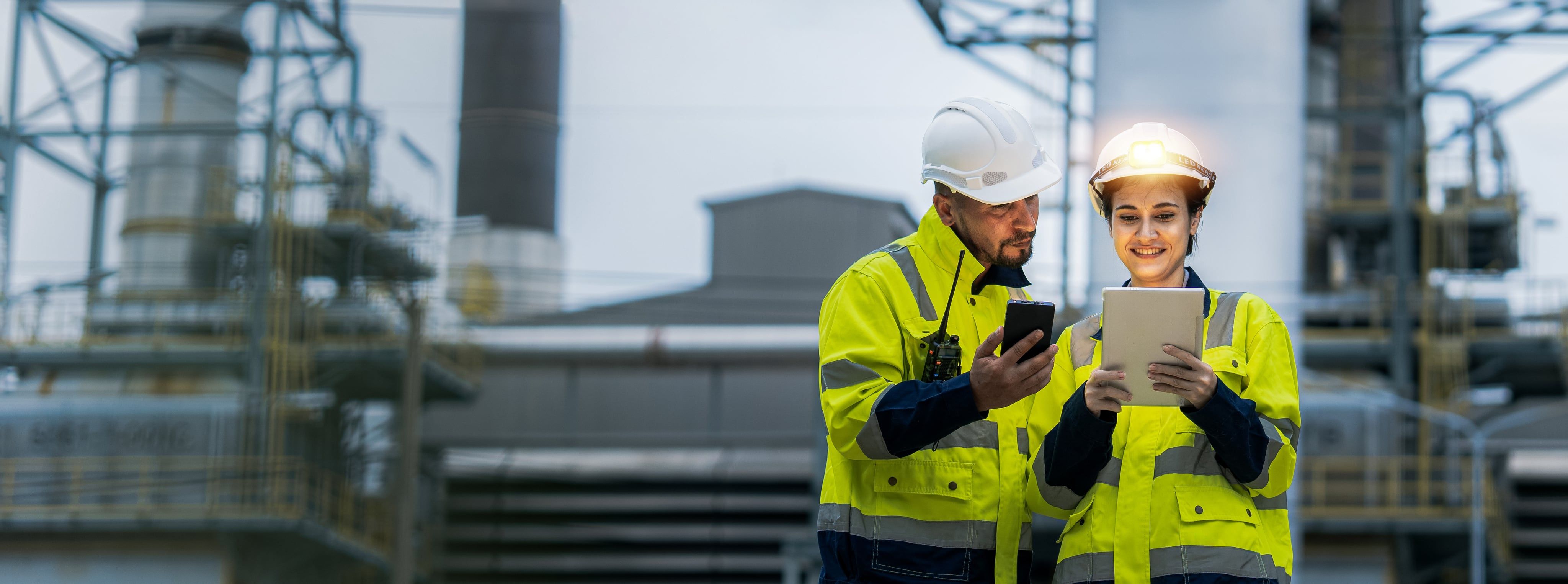 Two engineers in hard hats and high-visibility jackets checking energy data on a phone and a tablet at a plant.