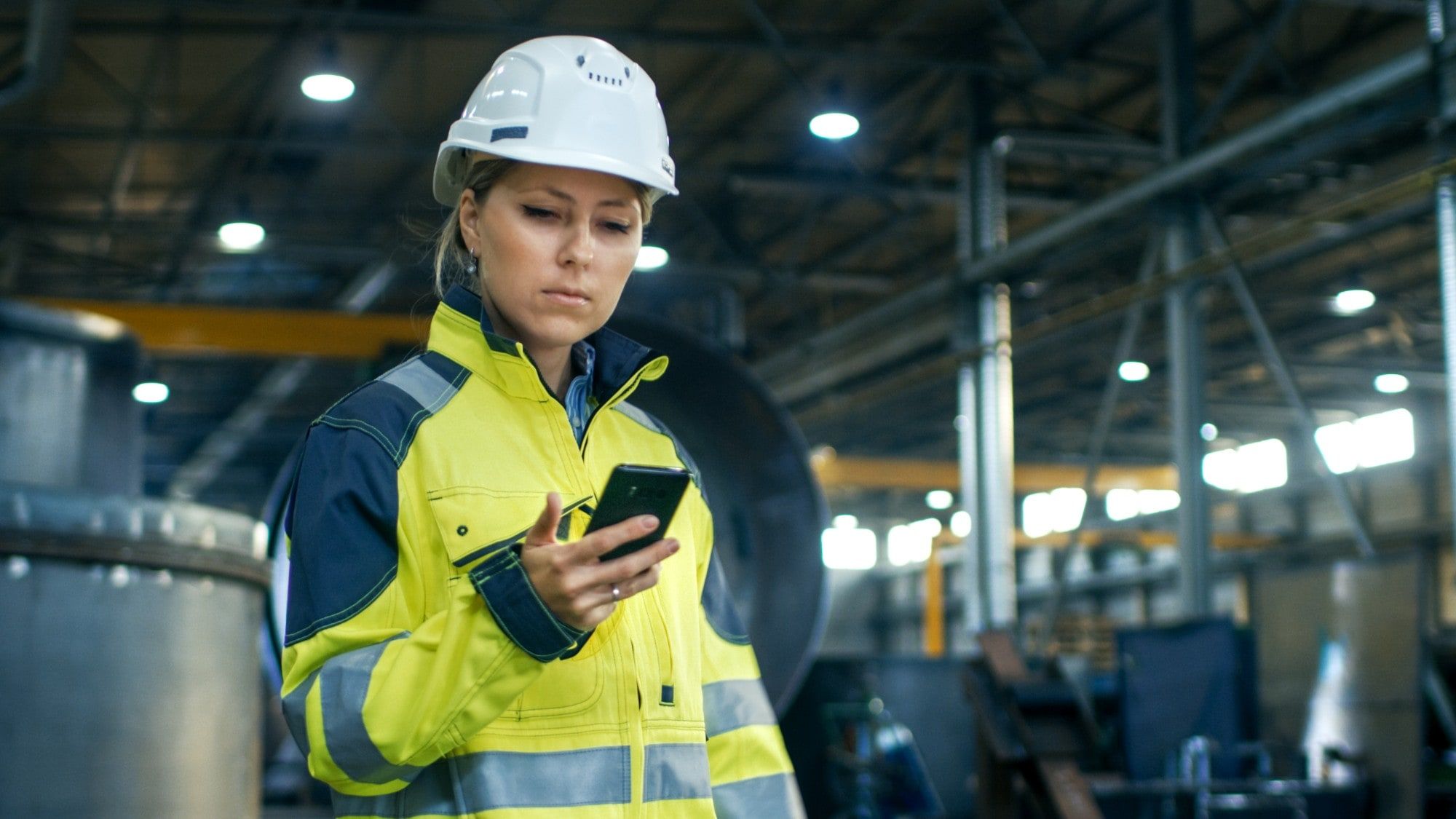 Engineer in a hard hat and high-visibility jacket checking energy data on her phone in a factory hall.